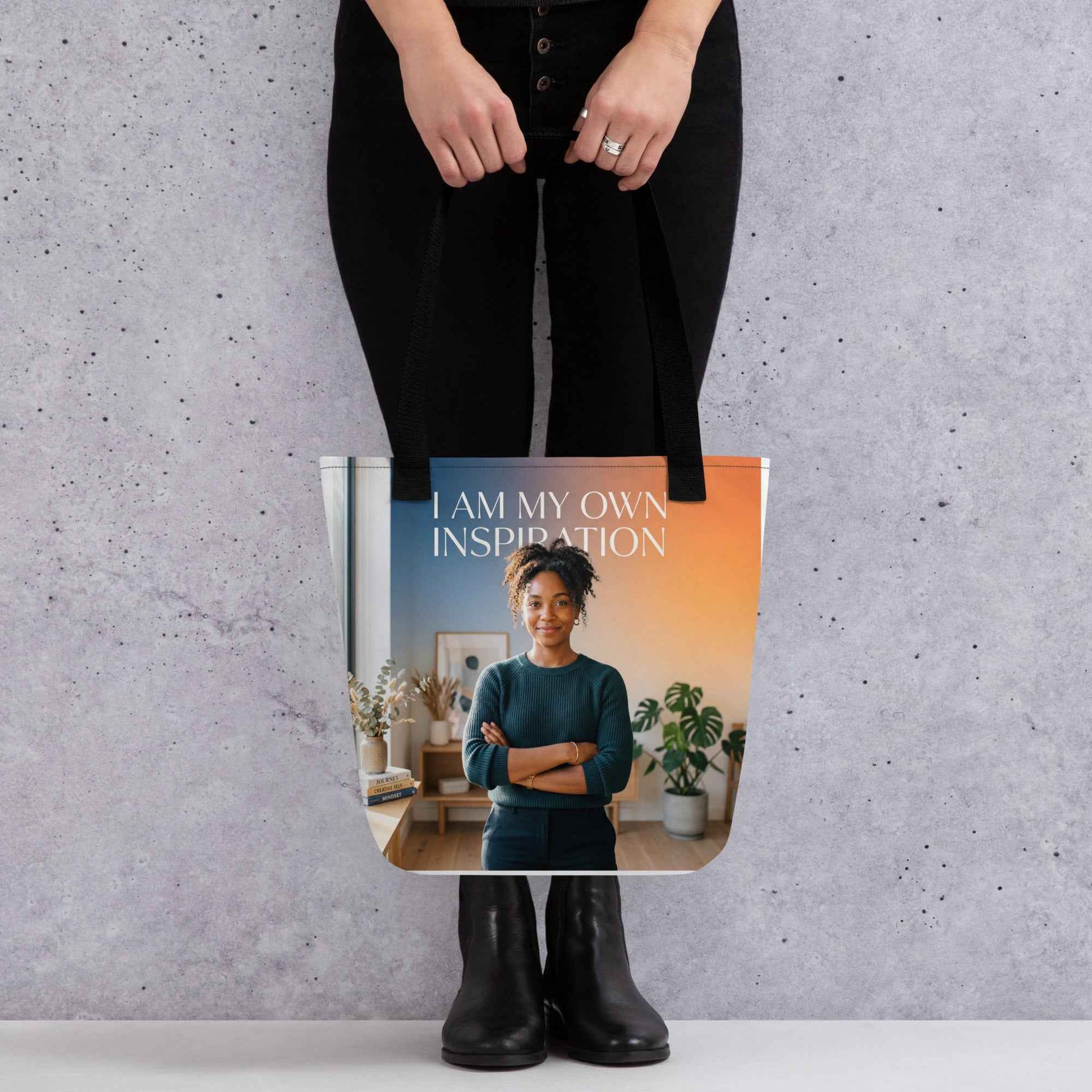 Printed tote bag held by black-clad person; “I AM MY OWN INSPIRATION” text and portrait.