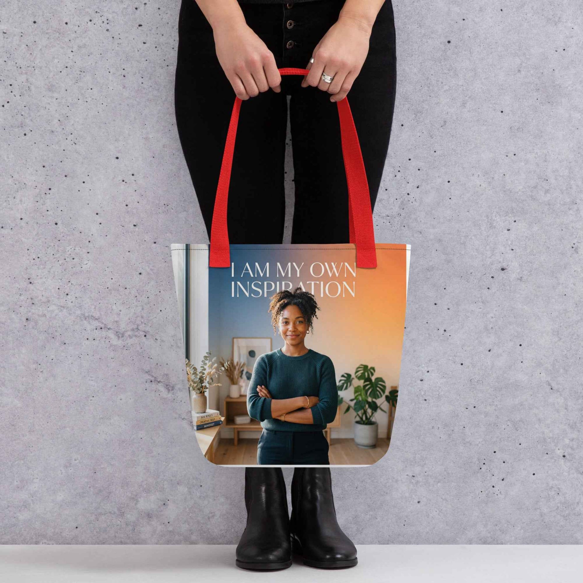 Printed tote bag held by black-clad person; red handles and “I AM MY OWN INSPIRATION” text.