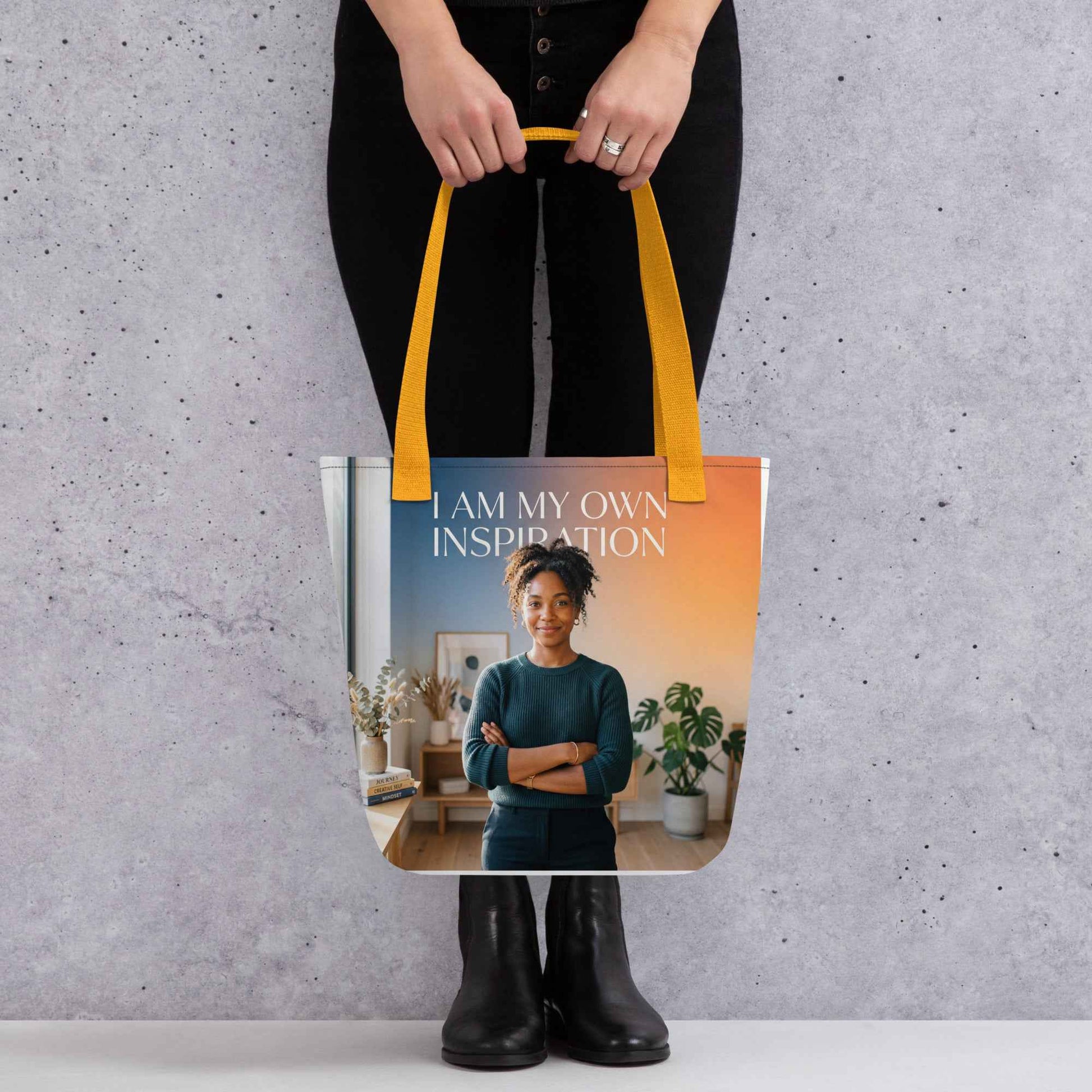 Printed tote bag held by black-clad person; yellow handles and “I AM MY OWN INSPIRATION” text.